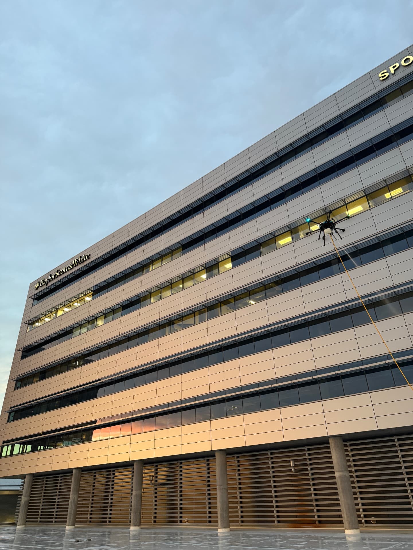 Cleaning drone working a hospital facade with fed water line at dusk