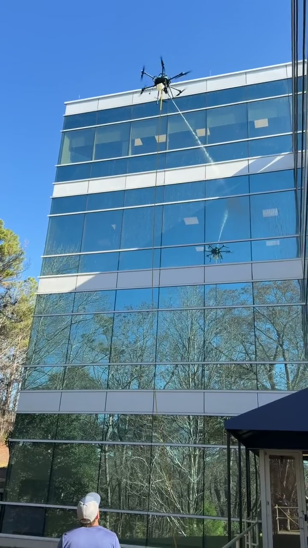 Drone washing the glass facade of a five-story office building while an operator watches from the ground